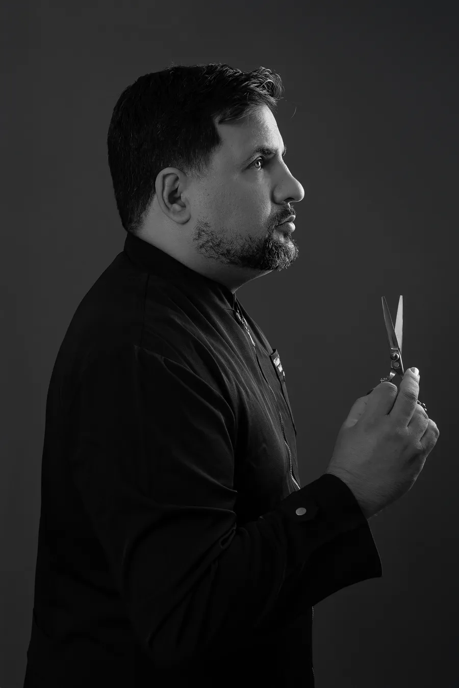 Verlano Alves in profile, holding shears, wearing black against a dark background